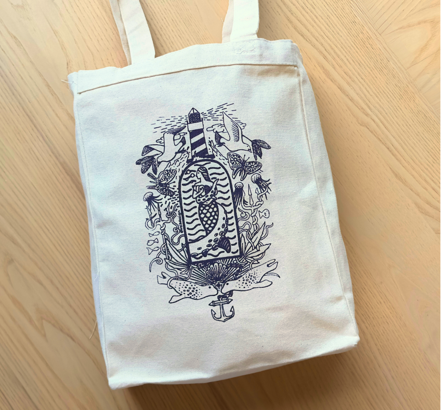 The SeaWeed Co. Canvas Tote - Purple is perfect for Maine adventures, featuring an illustrated lighthouse, waves, and sea creatures on cream-colored canvas. Resting on a light wooden floor, it beautifully captures a nautical theme.