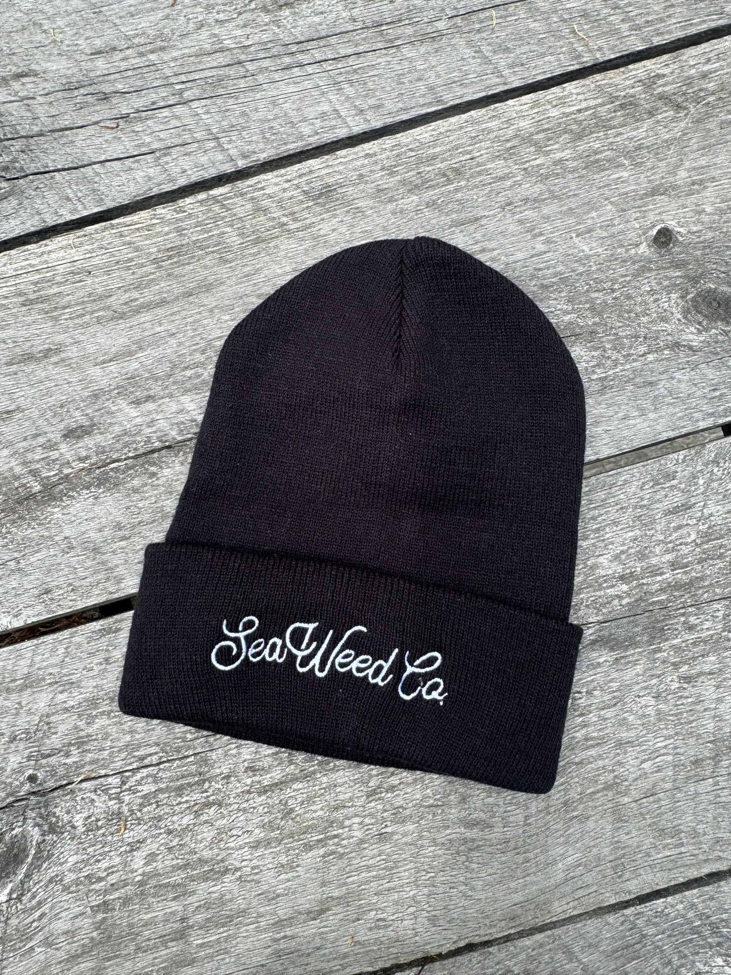 A cozy black Embroidered Beanie from SeaWeed Co., featuring elegant white embroidery, rests on a weathered wooden surface.