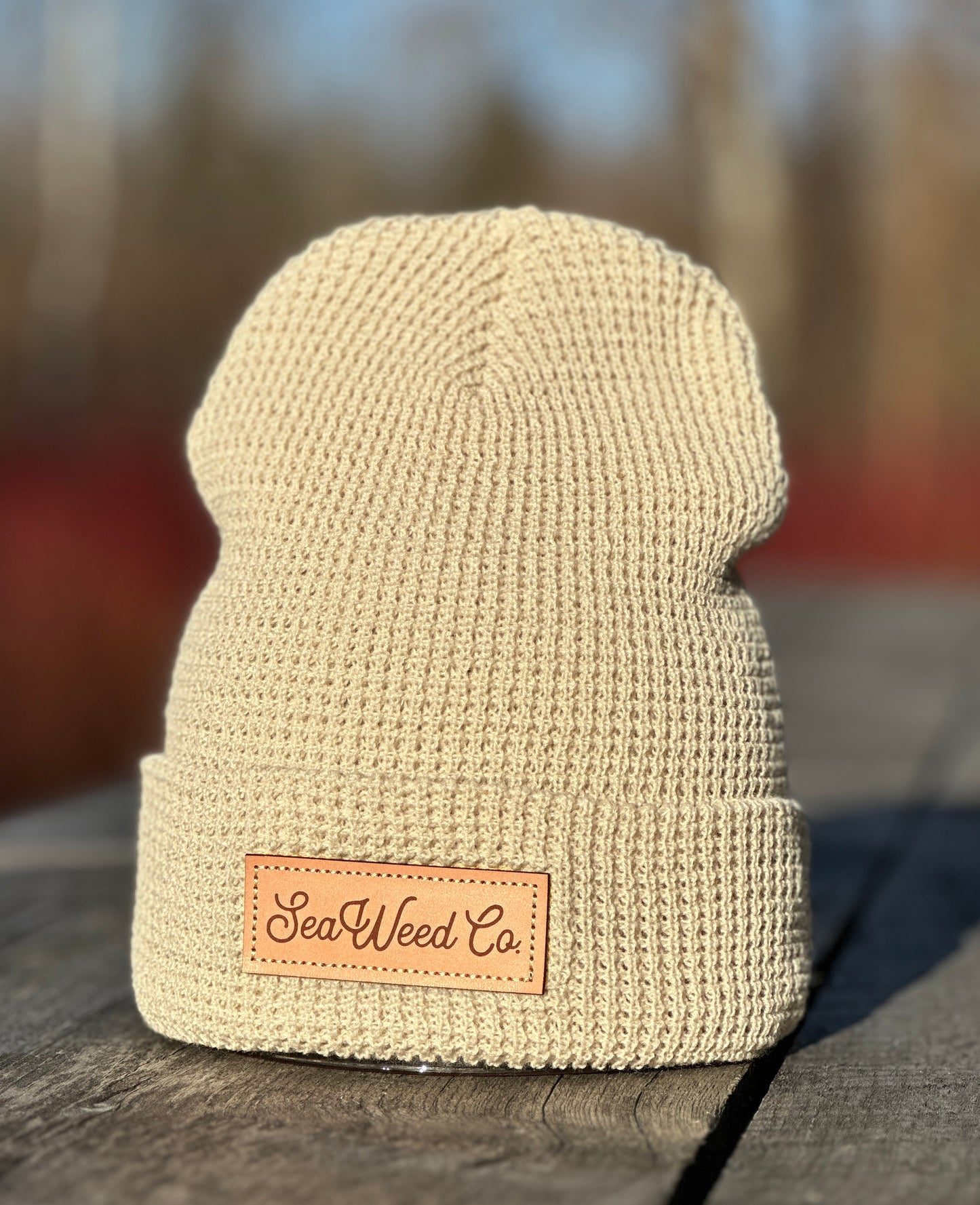 A close-up of the lightweight Beanie by SeaWeed Co. rests on a wooden table outdoors, featuring an embossed leather patch. The blurred background accentuates the knit fabrics texture.