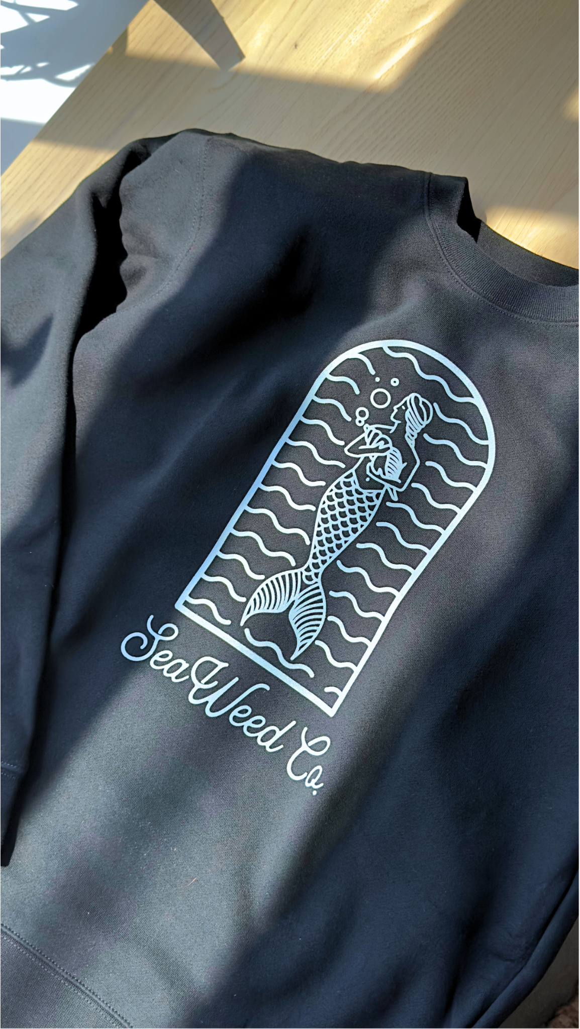 The SeaWeed Co. Mermaid Arch Crewneck - Black features a mermaid graphic with wavy lines in an arch. Made from soft cotton, it reads SeaWeed Co. below the illustration and is styled on a wooden surface with sunlight casting gentle shadows.