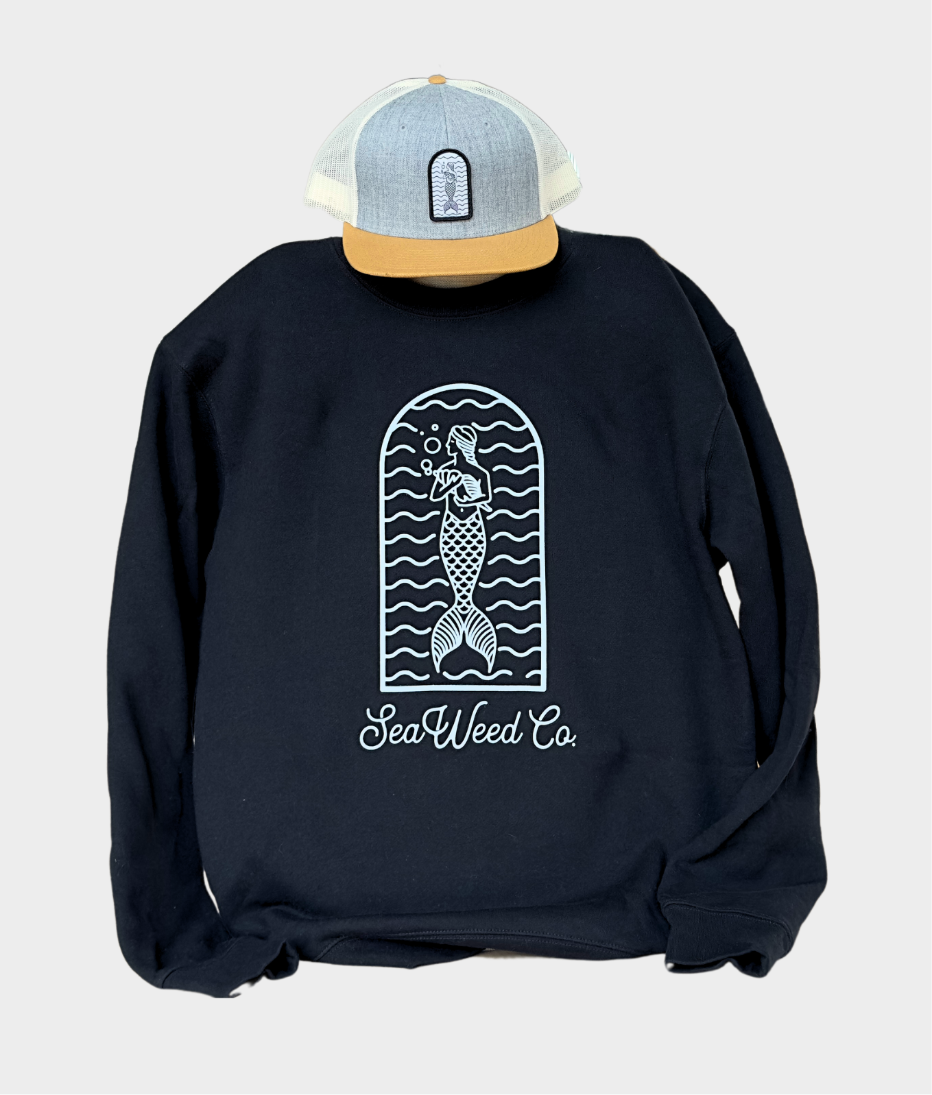 The SeaWeed Co. Mermaid Arch Crewneck features a black cotton sweatshirt with a white mermaid design and text SeaWeed Co. Paired with a gray/tan polyester trucker hat featuring the matching mermaid logo, this ensemble is the epitome of comfort and style.