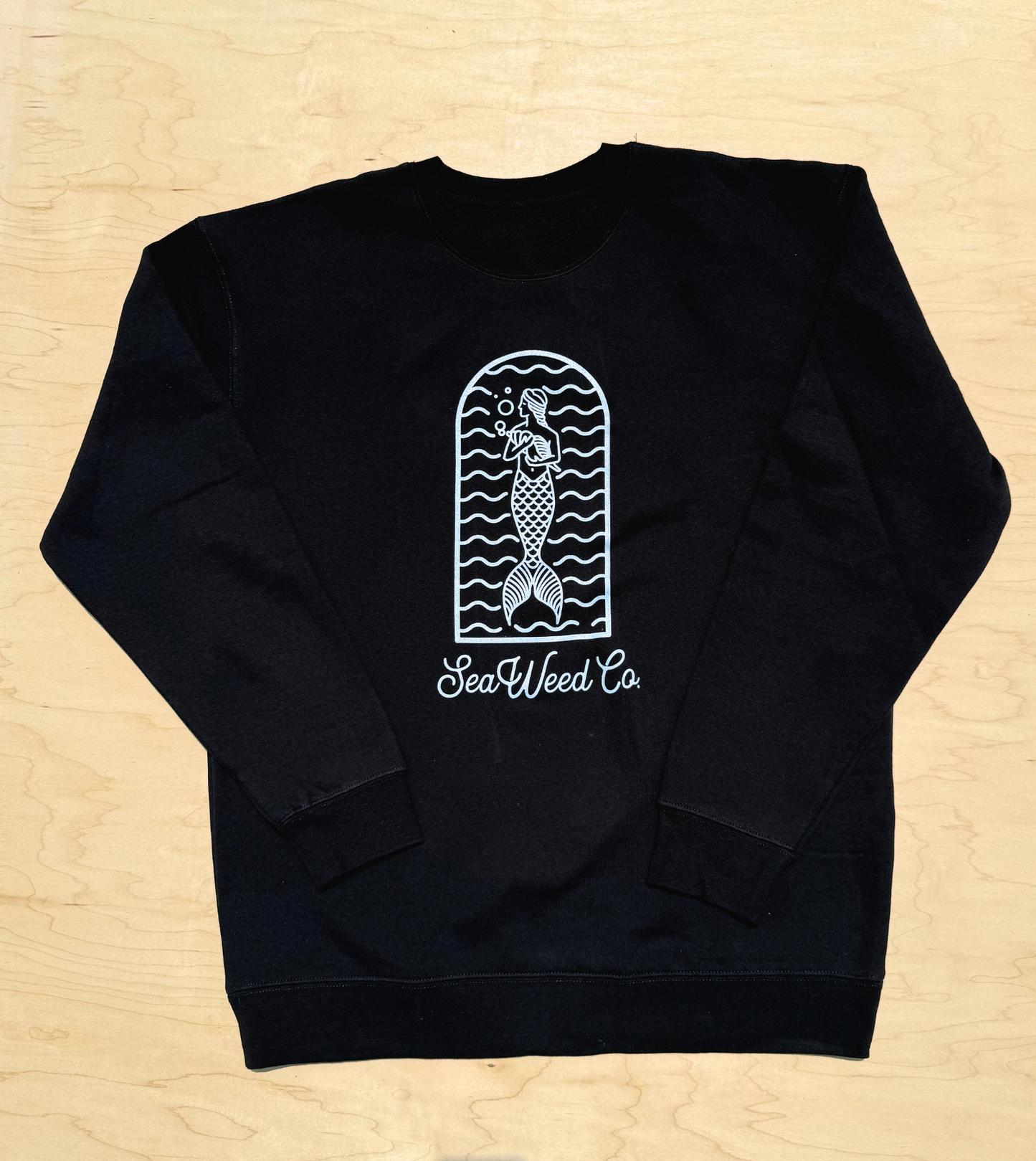 The Mermaid Arch Crewneck - Black from SeaWeed Co. features a white mermaid and wave design inside a window-like frame on a black polyester-cotton blend sweatshirt. Laid flat on light wood, it reads SeaWeed Co. and is part of the Seaweed Cozy Collection.