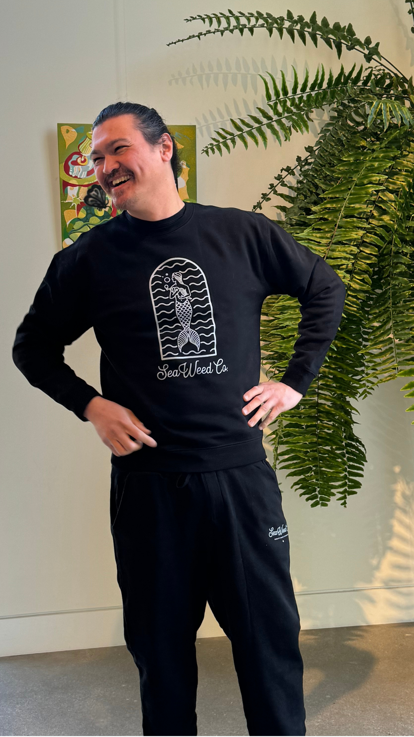 A person wearing a black Mermaid Arch Crewneck from SeaWeed Co. smiles while standing near a tall plant, with art visible on the wall behind them.