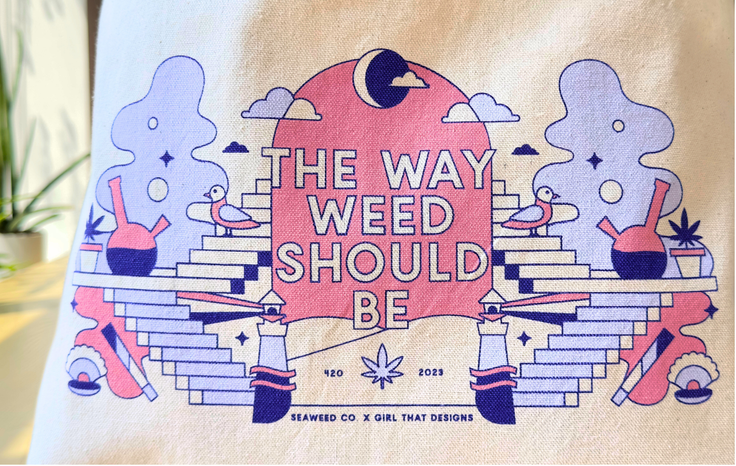 The Canvas Tote - TWWSB by SeaWeed Co. features a vibrant illustration with bongs, joints, and plants against a pink sunset with clouds and stars. It displays The Way Weed Should Be and is designed for Maine adventures by Seaweed Co. x Girl That Designs for 420 2023.