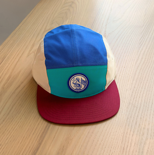 On a wooden table rests the SeaWeed Co. Colorblock Hat, displaying blue, teal, and beige panels with a striking red brim. The front features a circular patch with a Mermaid adding charm and playfulness.