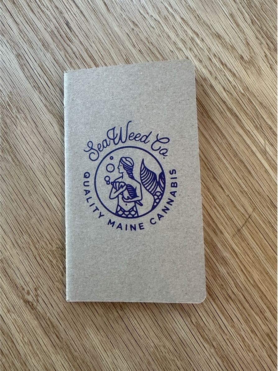 A notebook with a beige cover featuring the 'SeaWeed Co. Maine Cannabis' logo in a circular design.