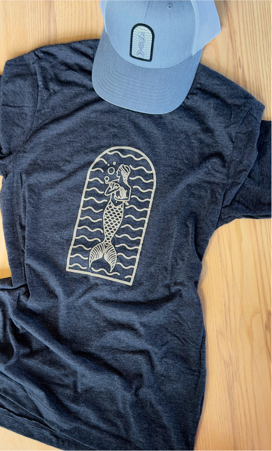 A gray t-shirt with a mermaid graphic design lying flat on a wooden surface, accompanied by a blue hat.