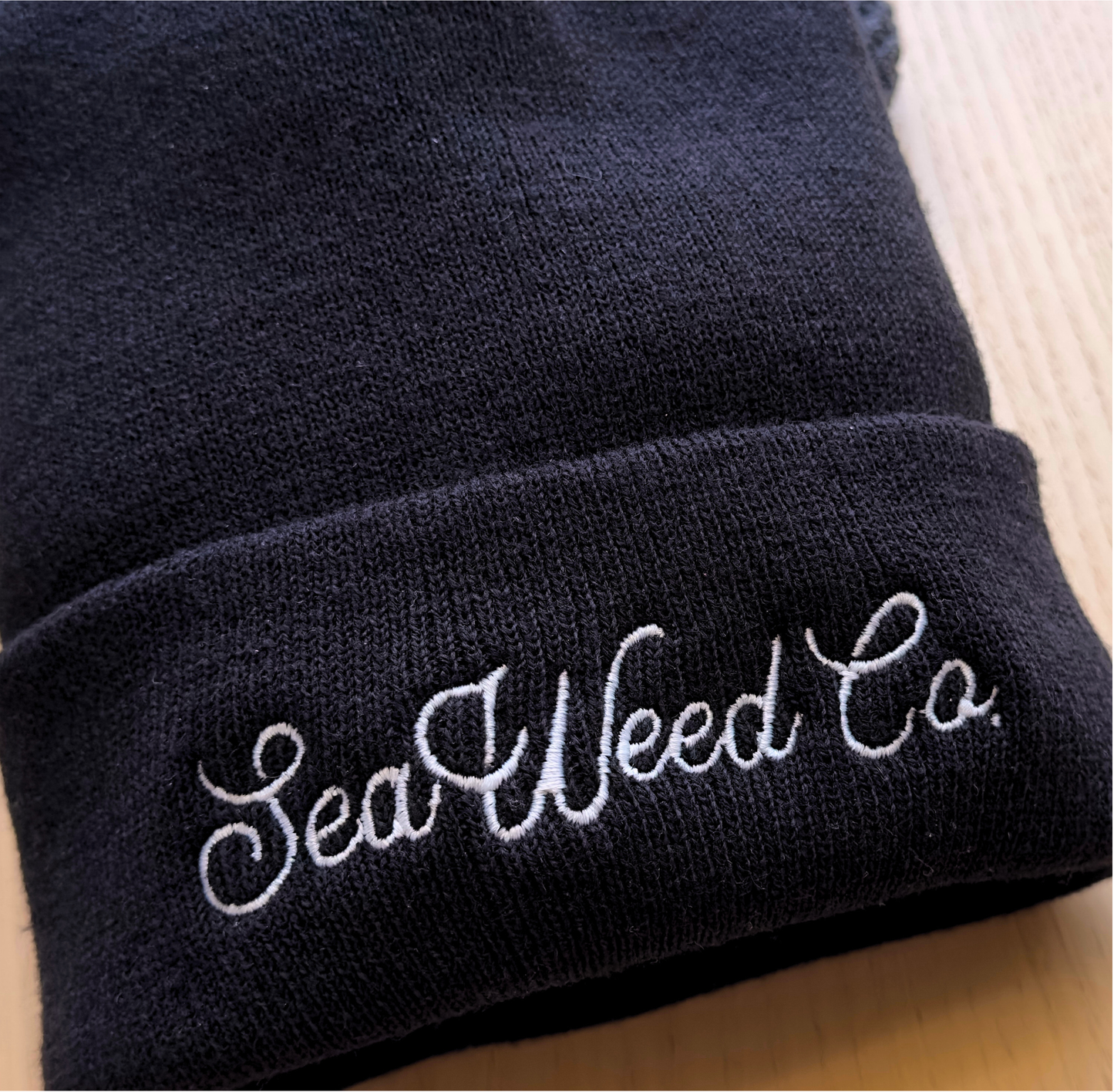 A cozy Embroidered Beanie in black, featuring SeaWeed Co. elegantly embroidered in white cursive on the folded brim, rests on a light wooden surface.